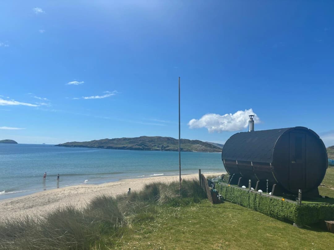 A dark barrel sauna sits on grass near a sandy beach and blue sea under a clear sky.