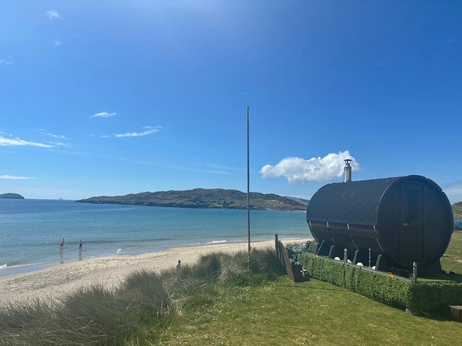 A dark barrel sauna sits on grass near a sandy beach and blue sea under a clear sky.
