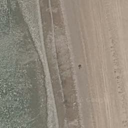 An overhead view shows a small black sauna structure on a sandy beach next to the sea.