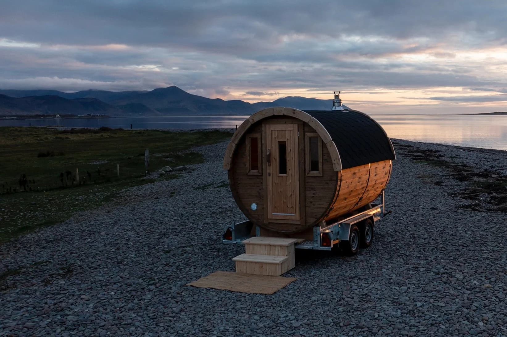A wooden barrel sauna on a trailer sits on a stony beach near the sea at dusk.