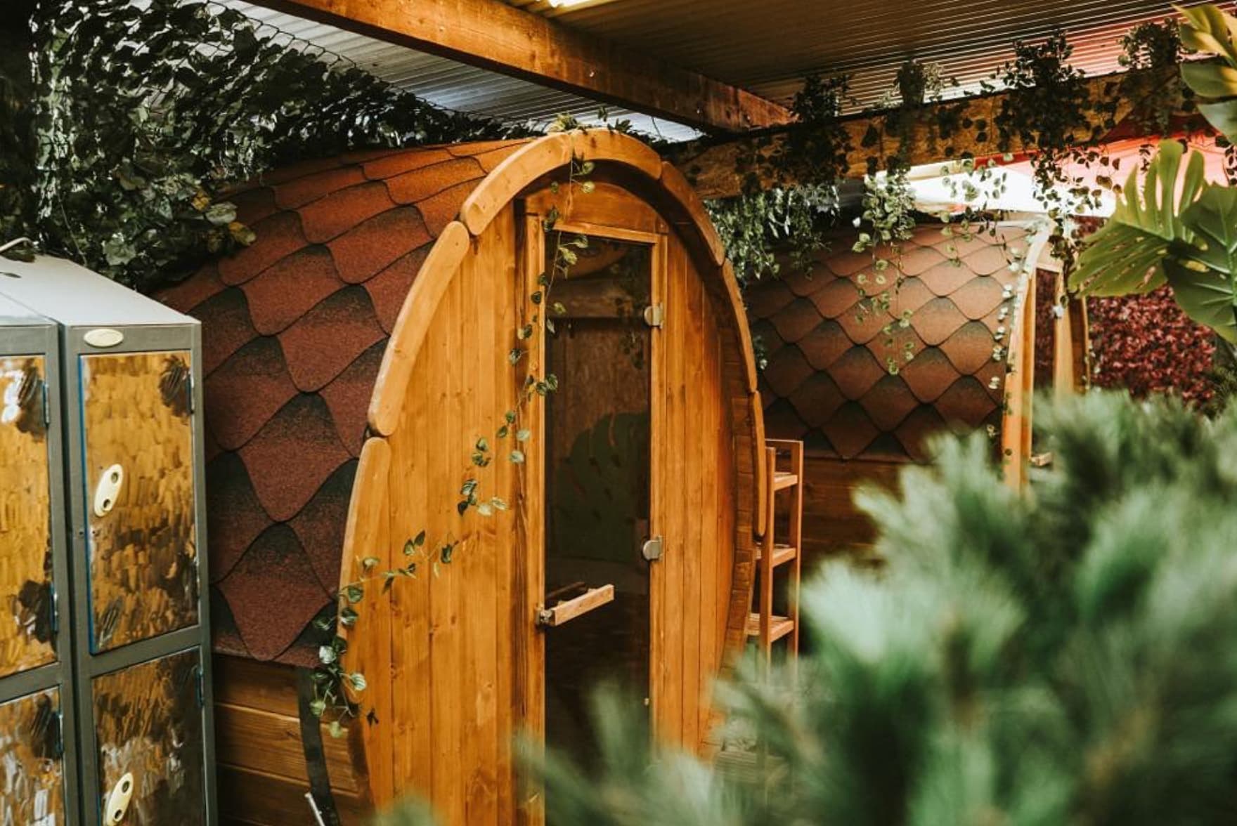 Wooden barrel saunas with shingled roofs are set indoors under a corrugated metal ceiling adorned with hanging plants.