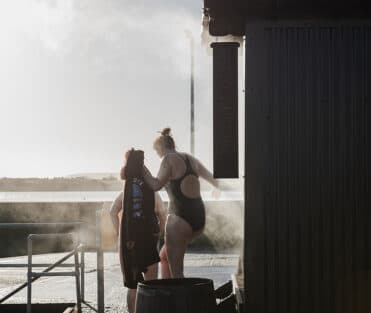People in swimwear near a dark sauna building with a vertical sign, overlooking a body of water.