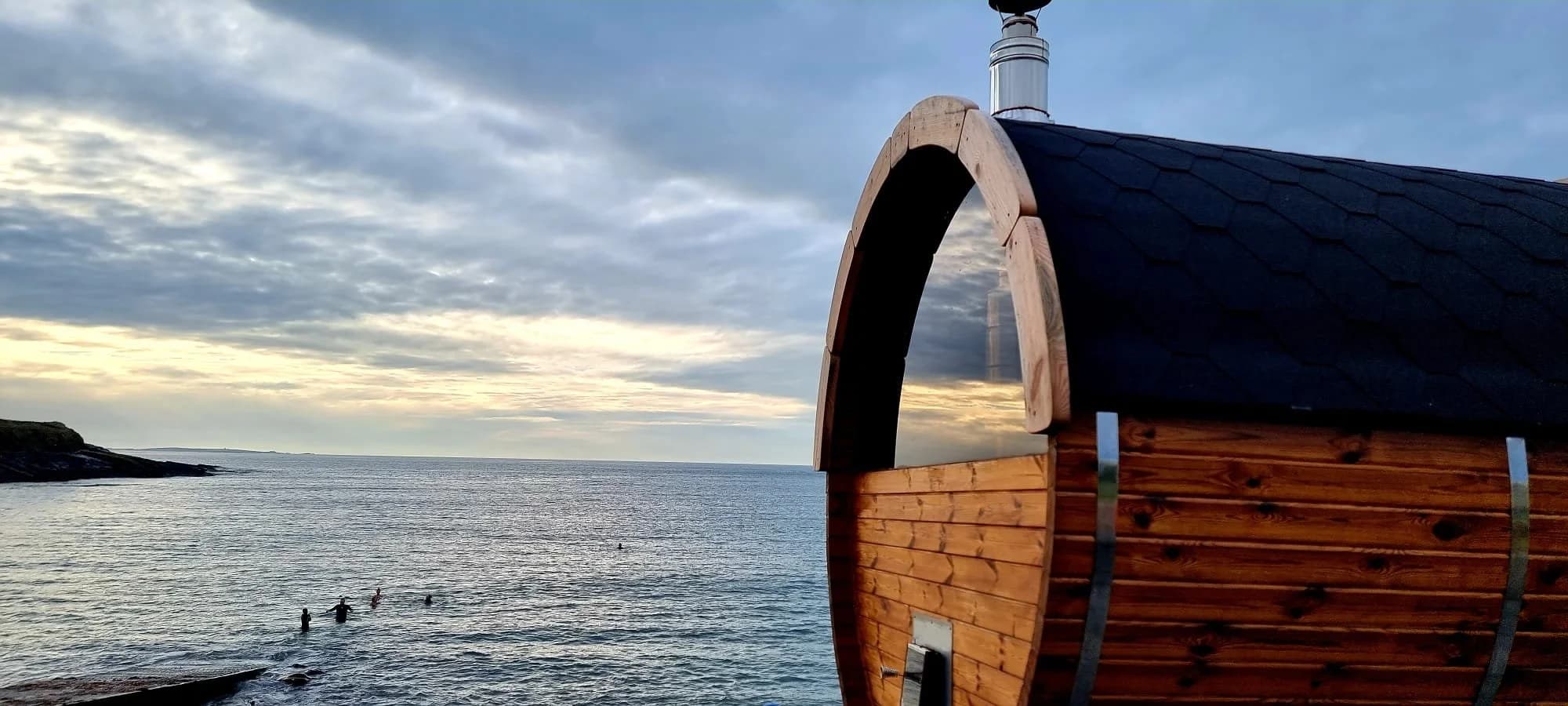 A wooden barrel sauna overlooks the ocean where people are swimming in the sea.