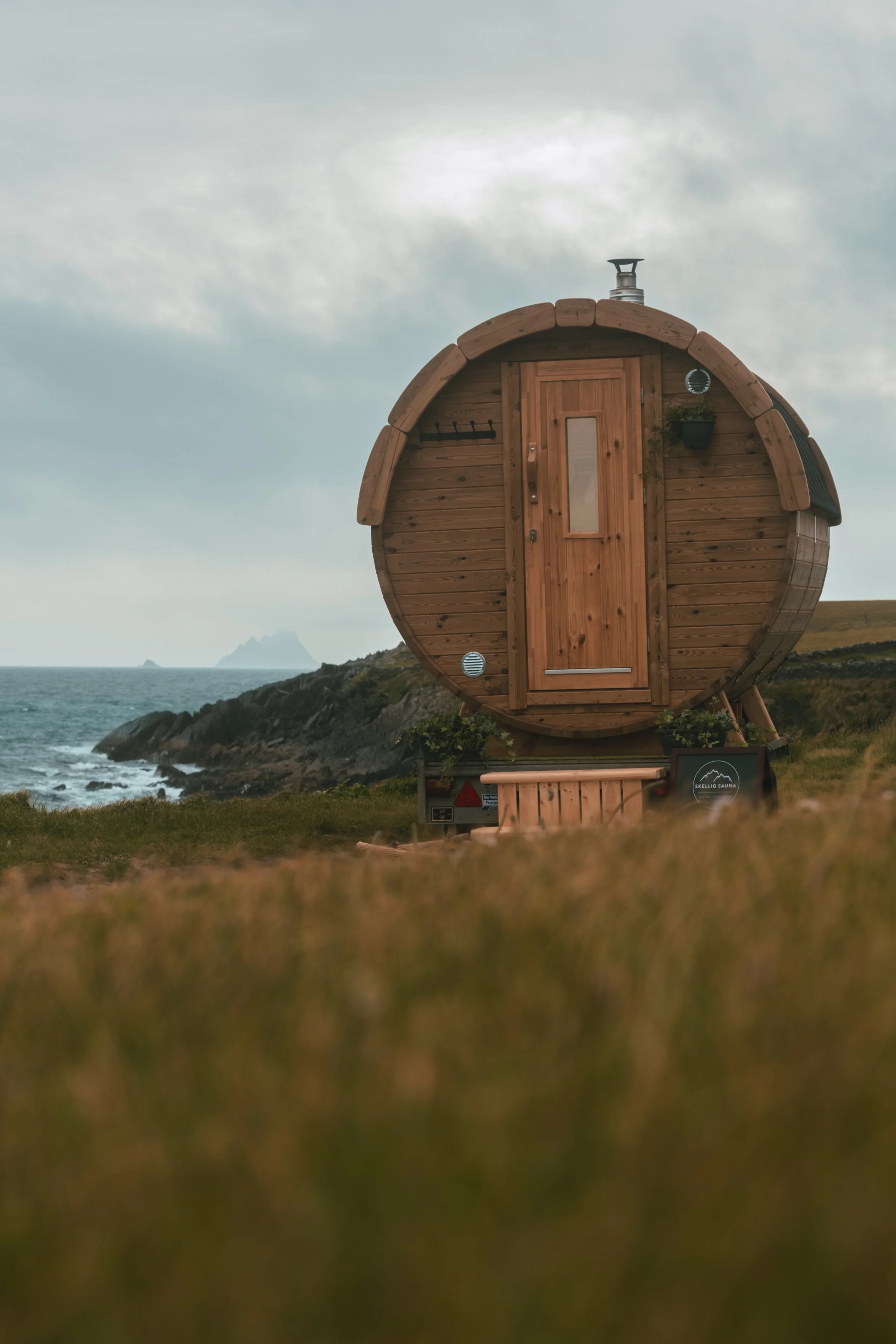 A barrel-shaped wooden sauna sits on a grassy cliff overlooking the ocean under an overcast sky.