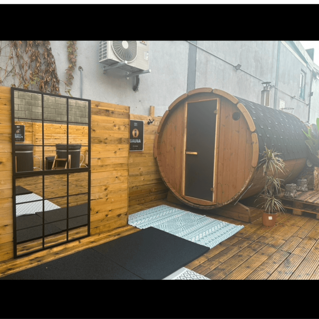 An outdoor barrel-shaped sauna on a wooden deck next to a privacy fence with a large rectangular mirror.