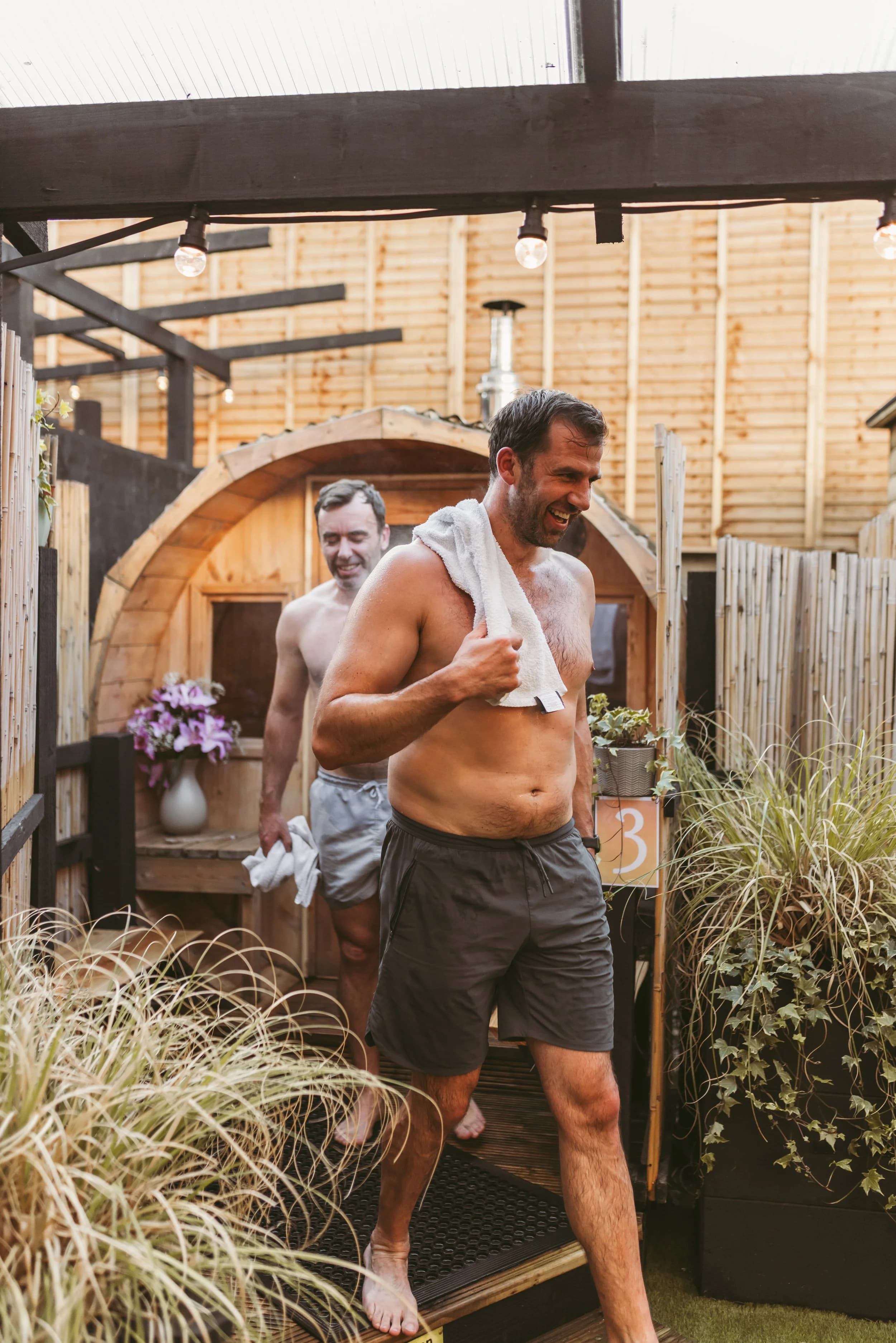 Two men leave a wooden barrel sauna in an outdoor setting, with one smiling and a towel on his shoulder.