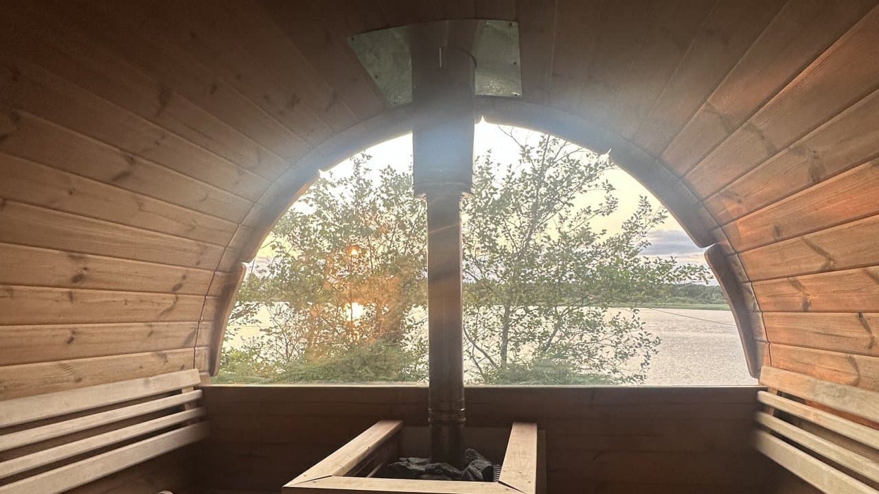 Wooden sauna interior with a stove and a large arched window framing trees, water, and the horizon.