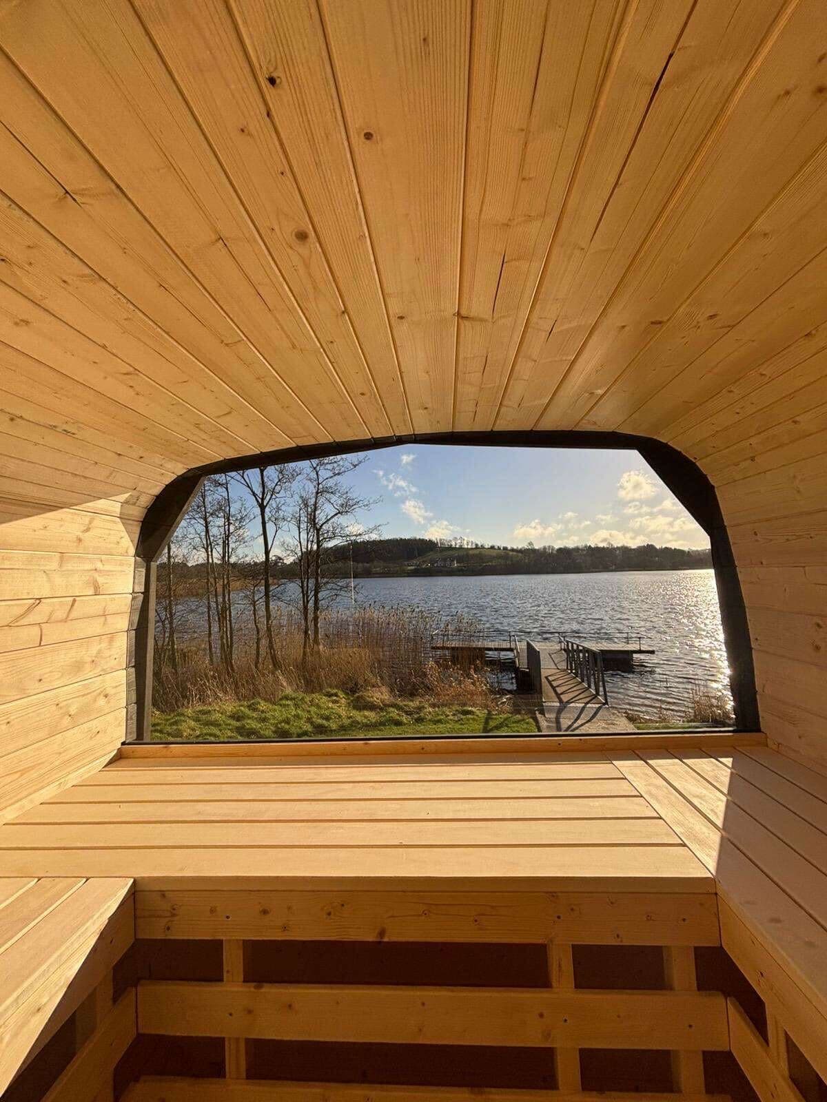 Interior view from a wooden sauna looking out through a large window toward a lake and dock.
